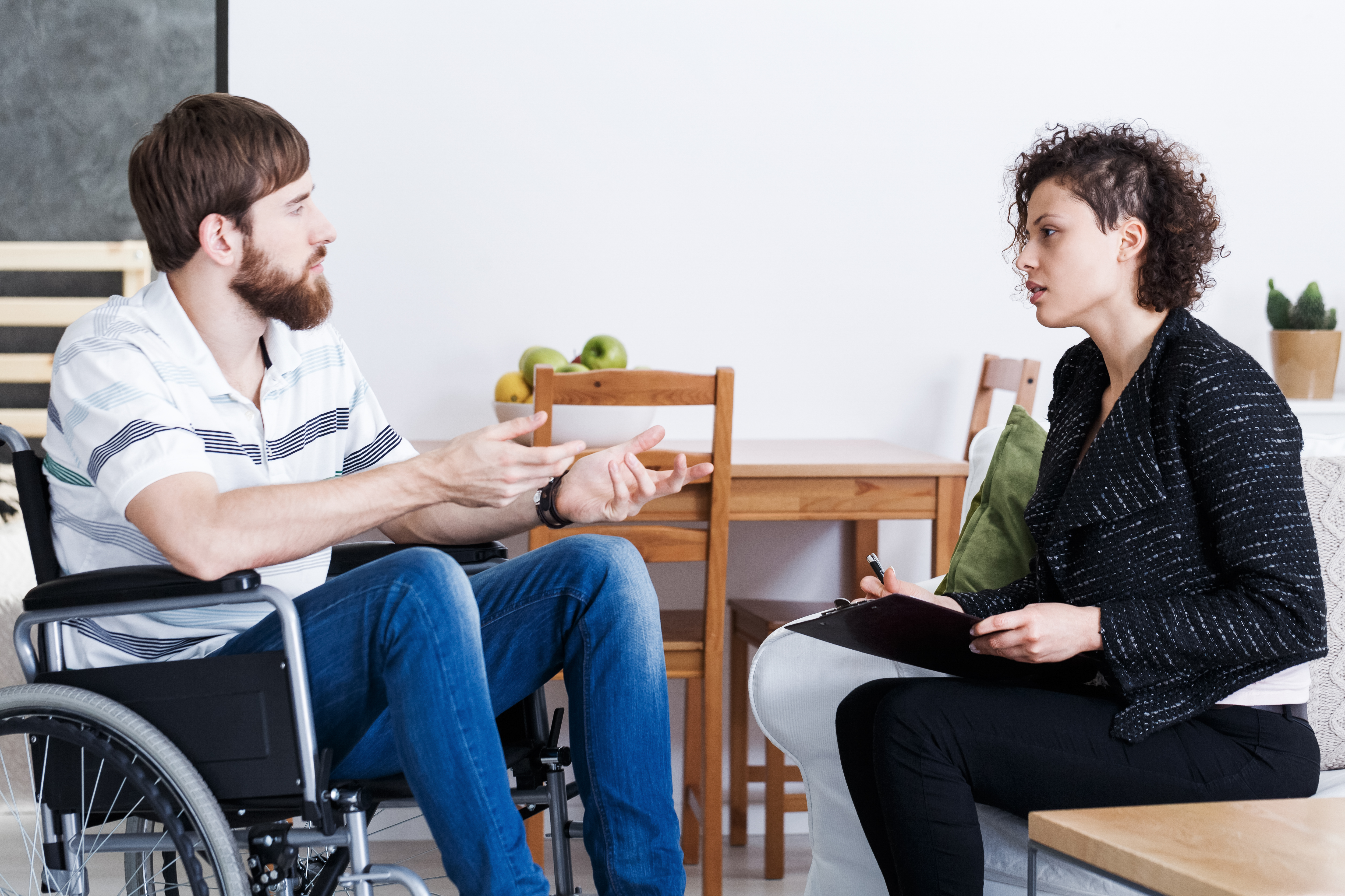 Young bearded man in wheelchair talking with psychotherapist at home